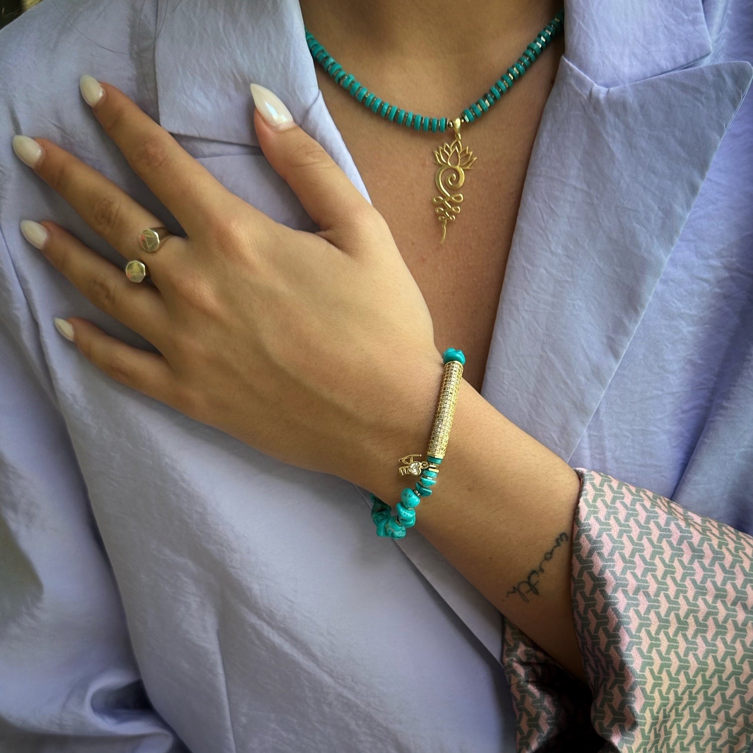 A hand model wearing the Diamond and Turquoise Love Bracelet, displaying its elegant design with turquoise stone beads, gold hematite spacers, and a gold plated love charm with a simulated diamond. The bracelet&#39;s stretchy jewelry cord ensures a comfortable fit.