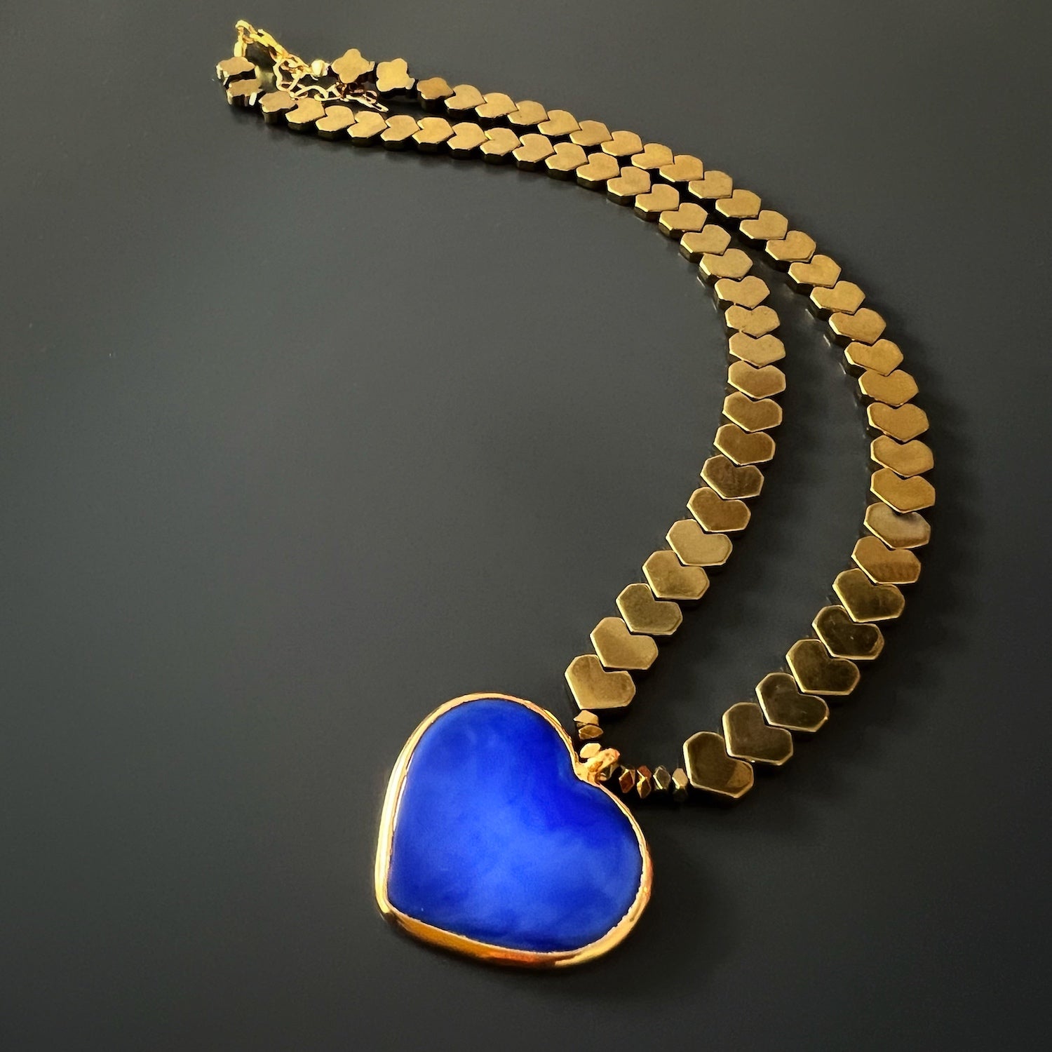 The Happy Blue Heart Necklace, displaying the arrangement of the gold color hematite beads and the blue heart ceramic pendant, illustrating the balanced and harmonious combination of colors and textures.
