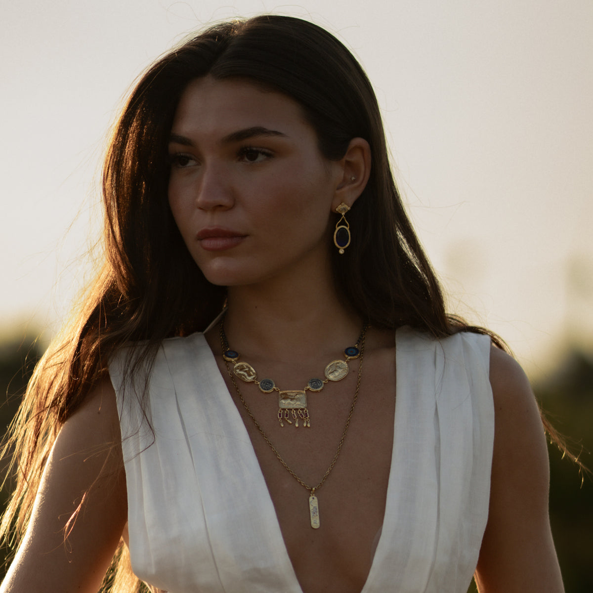 Woman in a white dress wearing the Amulet Gathering Evil Eye & Ruby Fringe Two Tone Necklace layered with a longer gold pendant