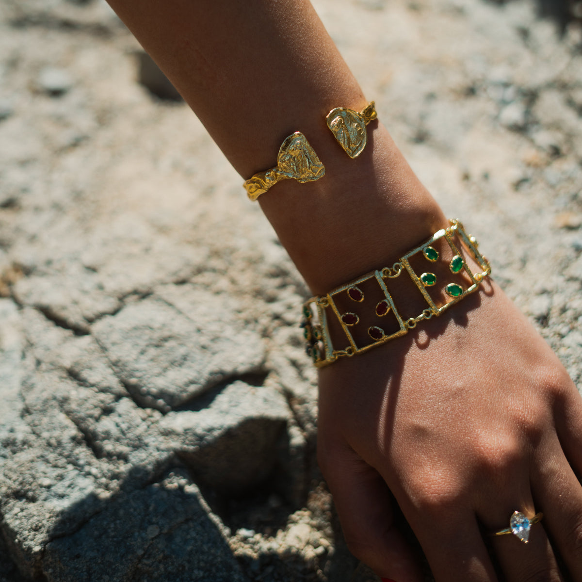 Hand resting on stone wearing the owl coin gold cuff, a jeweled link bracelet and a pear stone ring