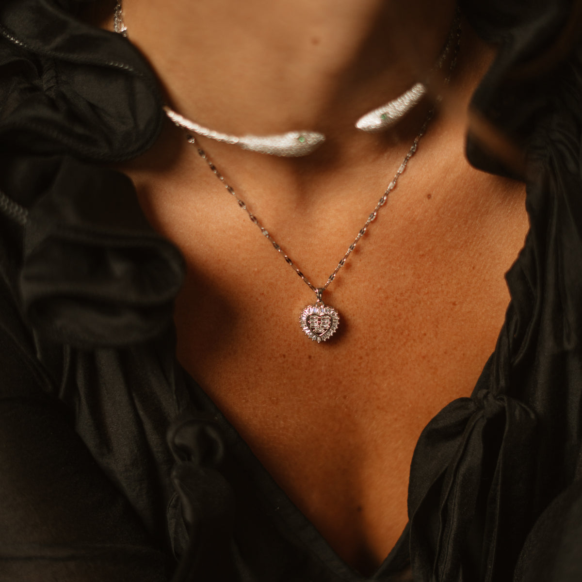 Close view of the baguette halo heart pendant at the neckline, layered under a silver collar with a black ruffled blouse