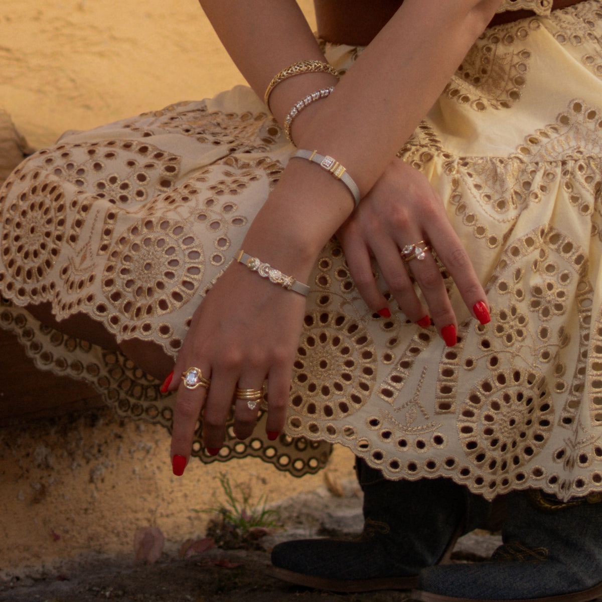 Model in a cream eyelet dress with a silver charm bracelet on each wrist, gold bangles and rings