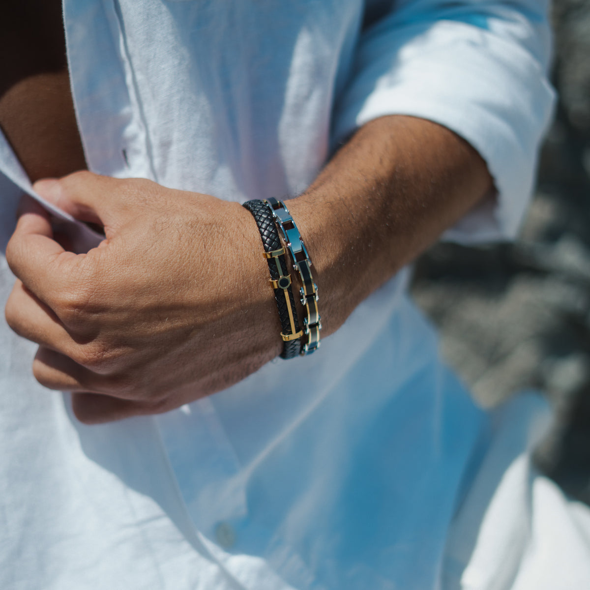 Black braided leather bracelet with a gold bar and black center accent on a man's wrist against a white shirt, paired with a blue and silver link bracelet