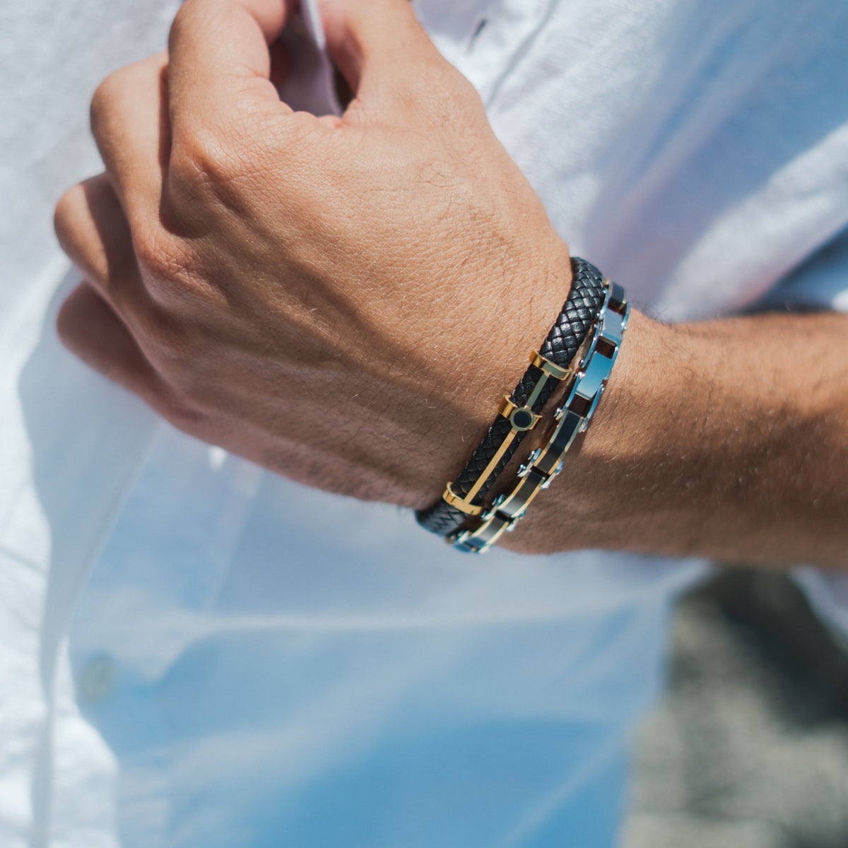 Man in a white linen shirt wearing the black braided leather gold bar men's bracelet stacked with a blue and silver link bracelet