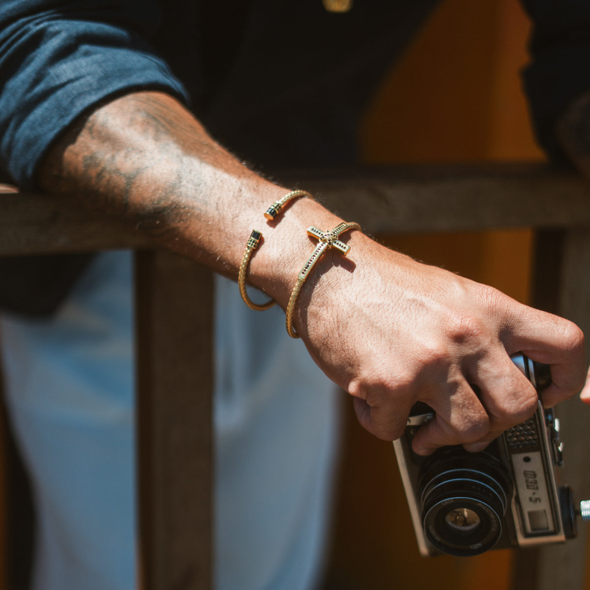Men's gold cross cuff bracelet worn with a thin gold cuff on a sunlit wrist, hand holding a camera
