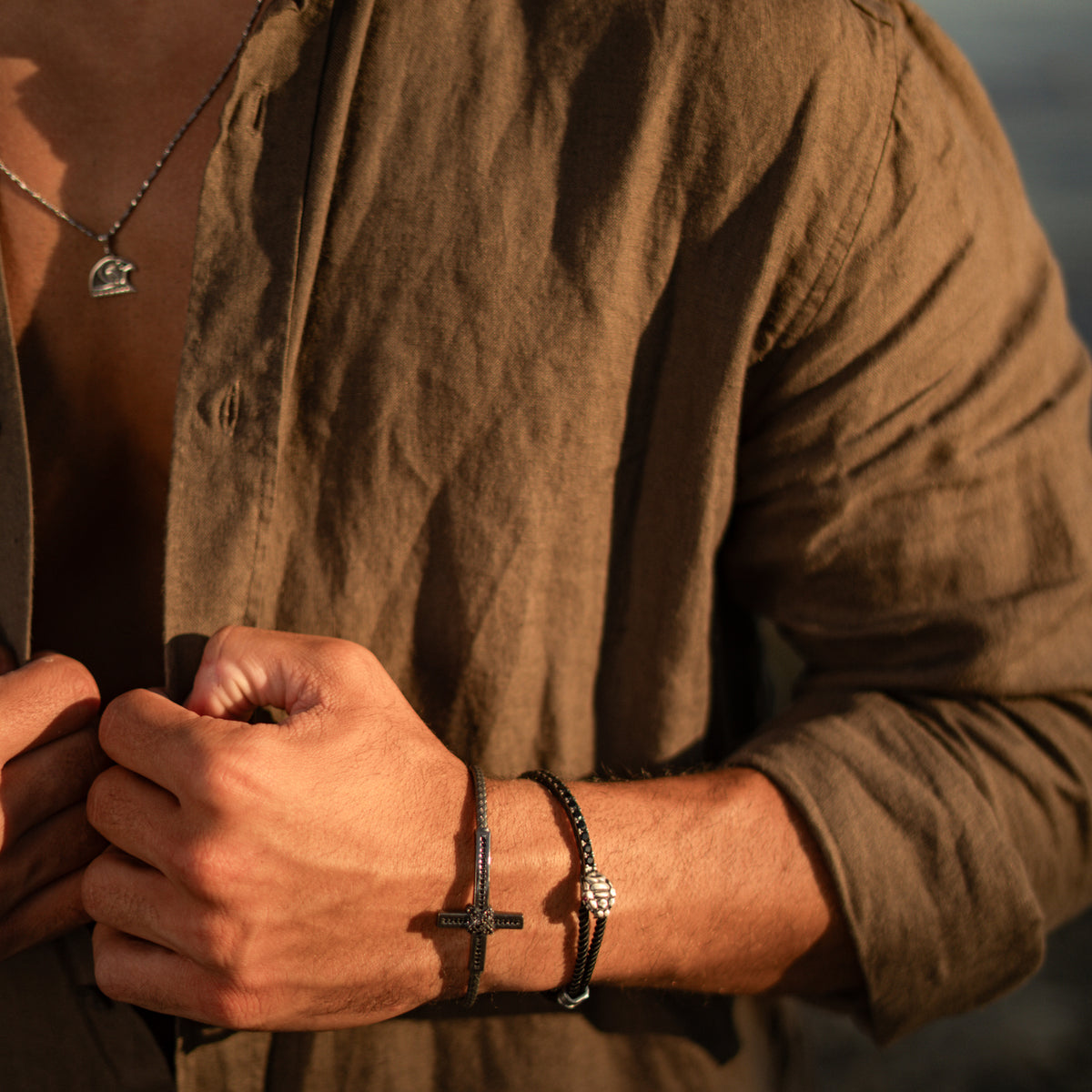 Man in an olive linen shirt and silver pendant necklace wearing the black zircon cross cuff bracelet in warm evening light