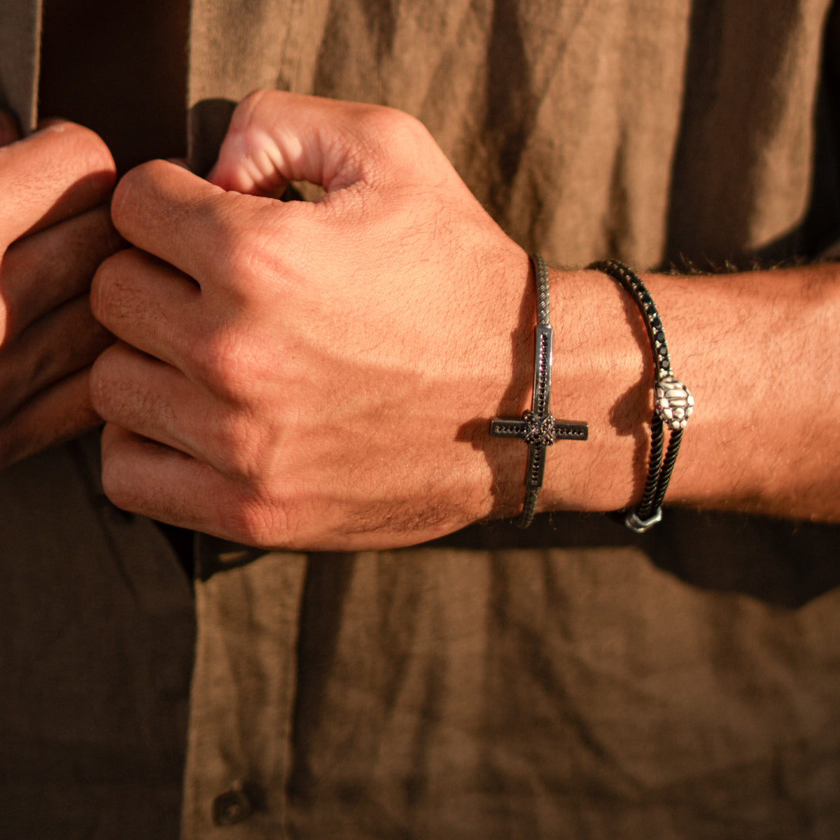 Man in a linen shirt wearing the oxidized silver cross cuff bracelet stacked with a black braided cord bracelet