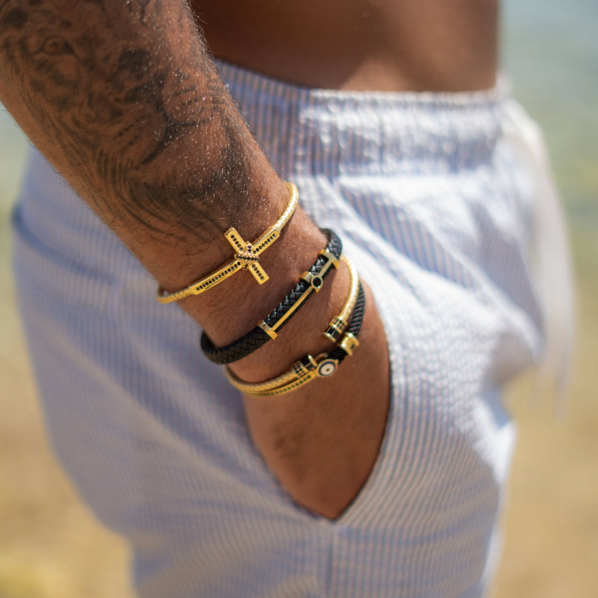 Stack of men's gold bracelets worn with a white striped shirt, including a gold cross cuff and a black braided leather gold bar bracelet