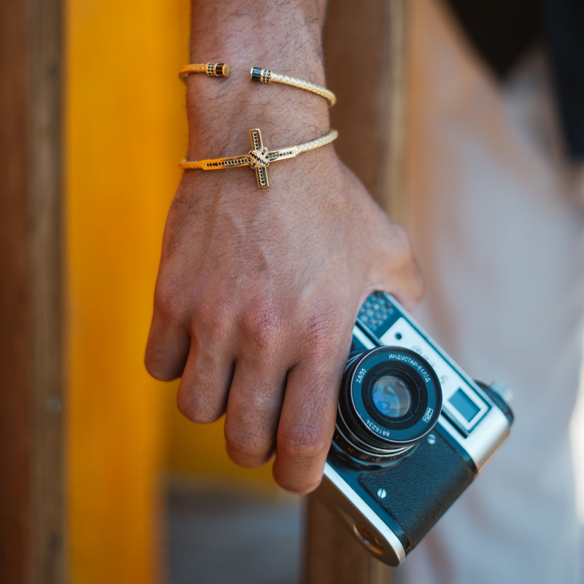 Man holding a vintage camera, wearing the slim woven gold vermeil cuff stacked with a gold cross cuff bracelet