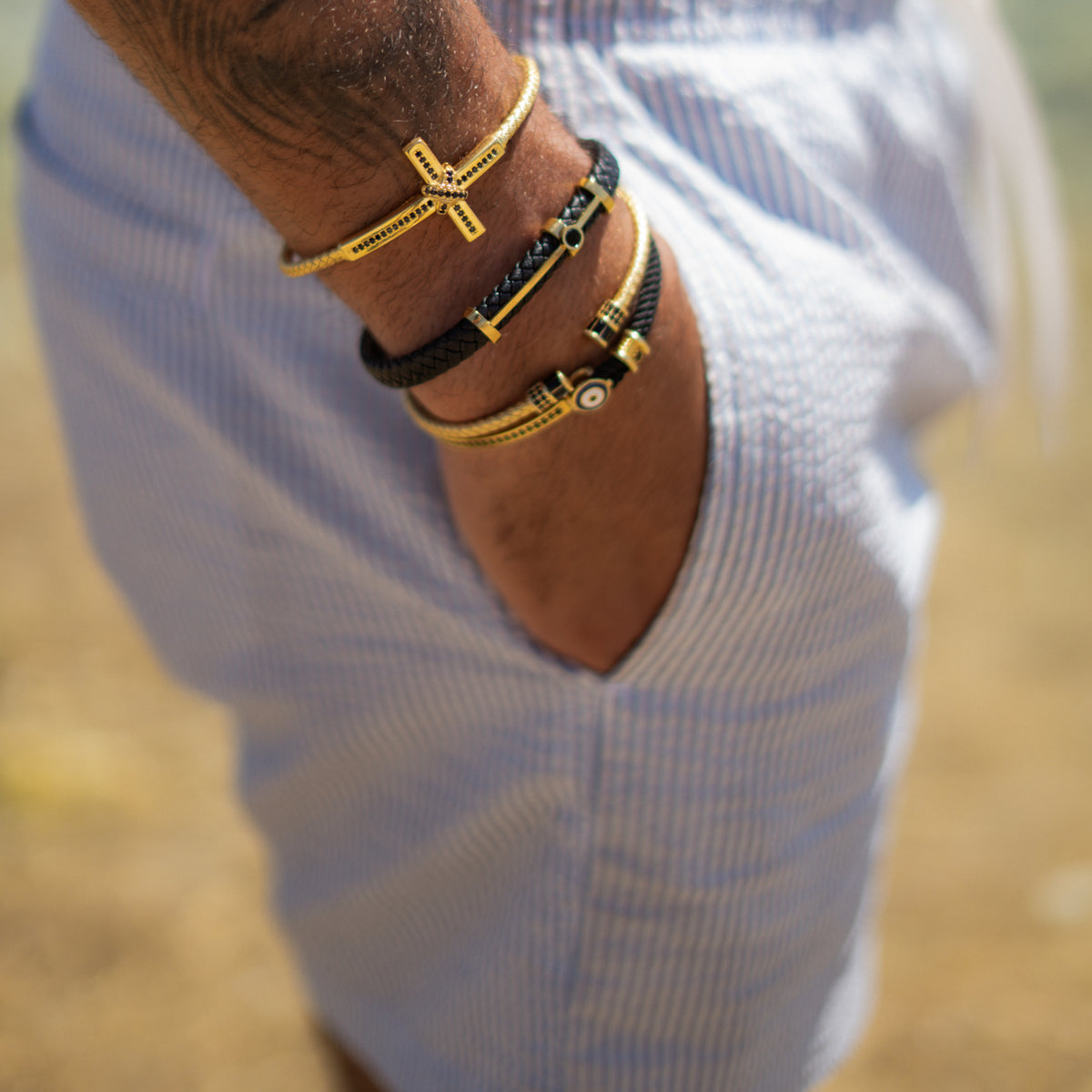 Black leather and gold bar men's bracelet stacked with gold cuffs on a wrist resting in a pocket, outdoors in sunlight