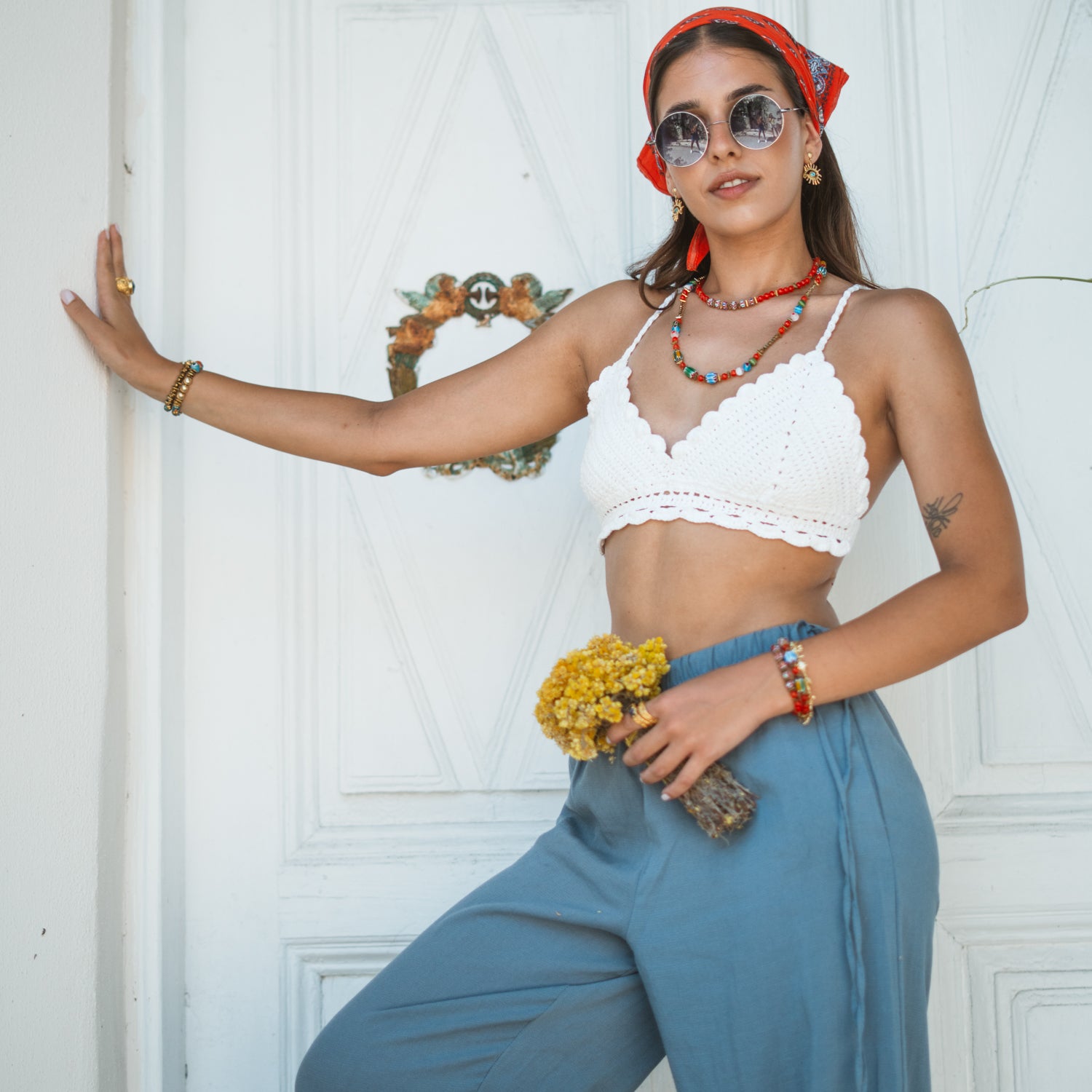 Model leaning against a white door wearing the handmade Carnelian Fire Energy Beaded Choker, a white crochet top, and blue linen pants