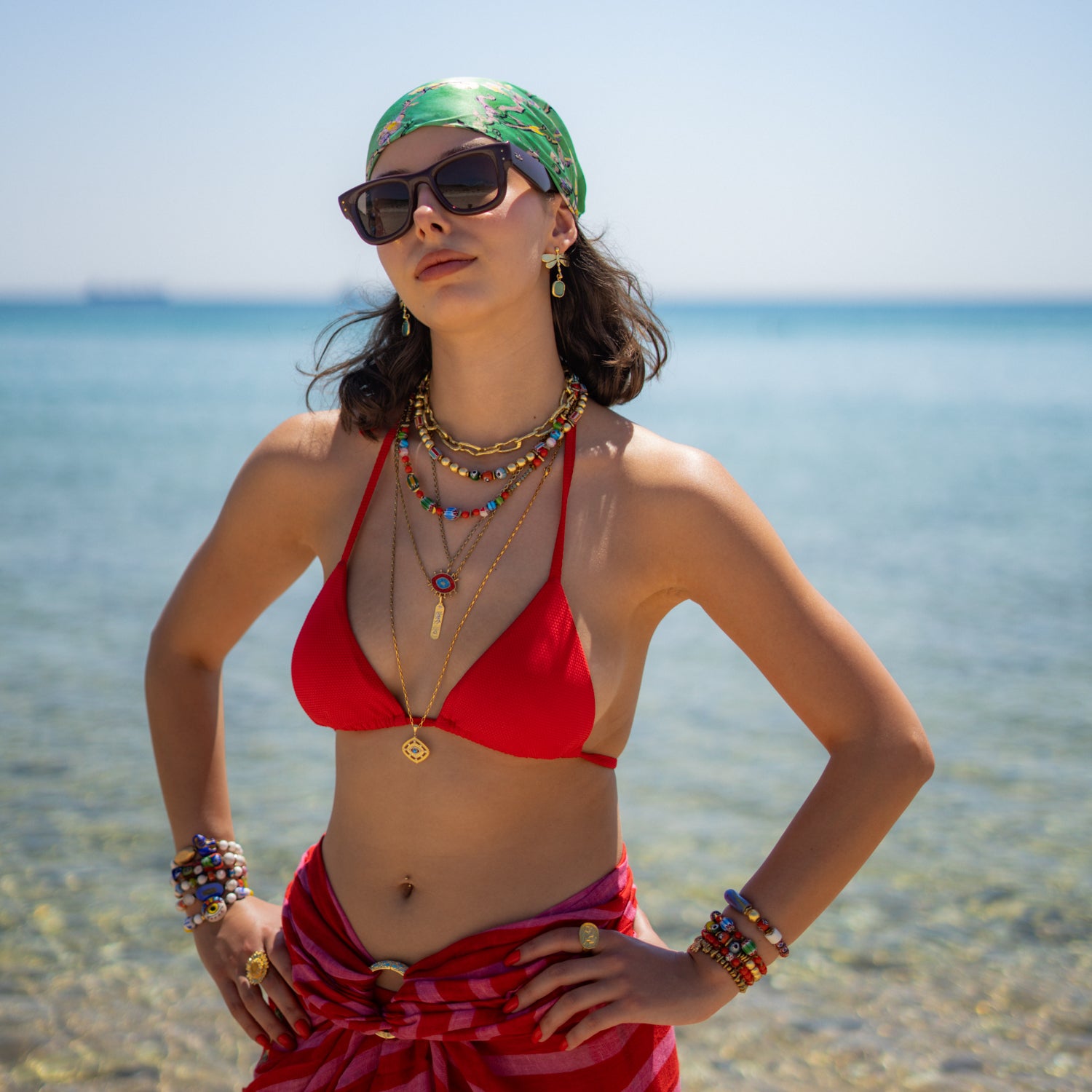 Model at the beach in a red bikini wearing the colorful boho Carnelian Rainbow Energy Beaded Necklace, layered gold necklaces, beaded bracelets, and rings