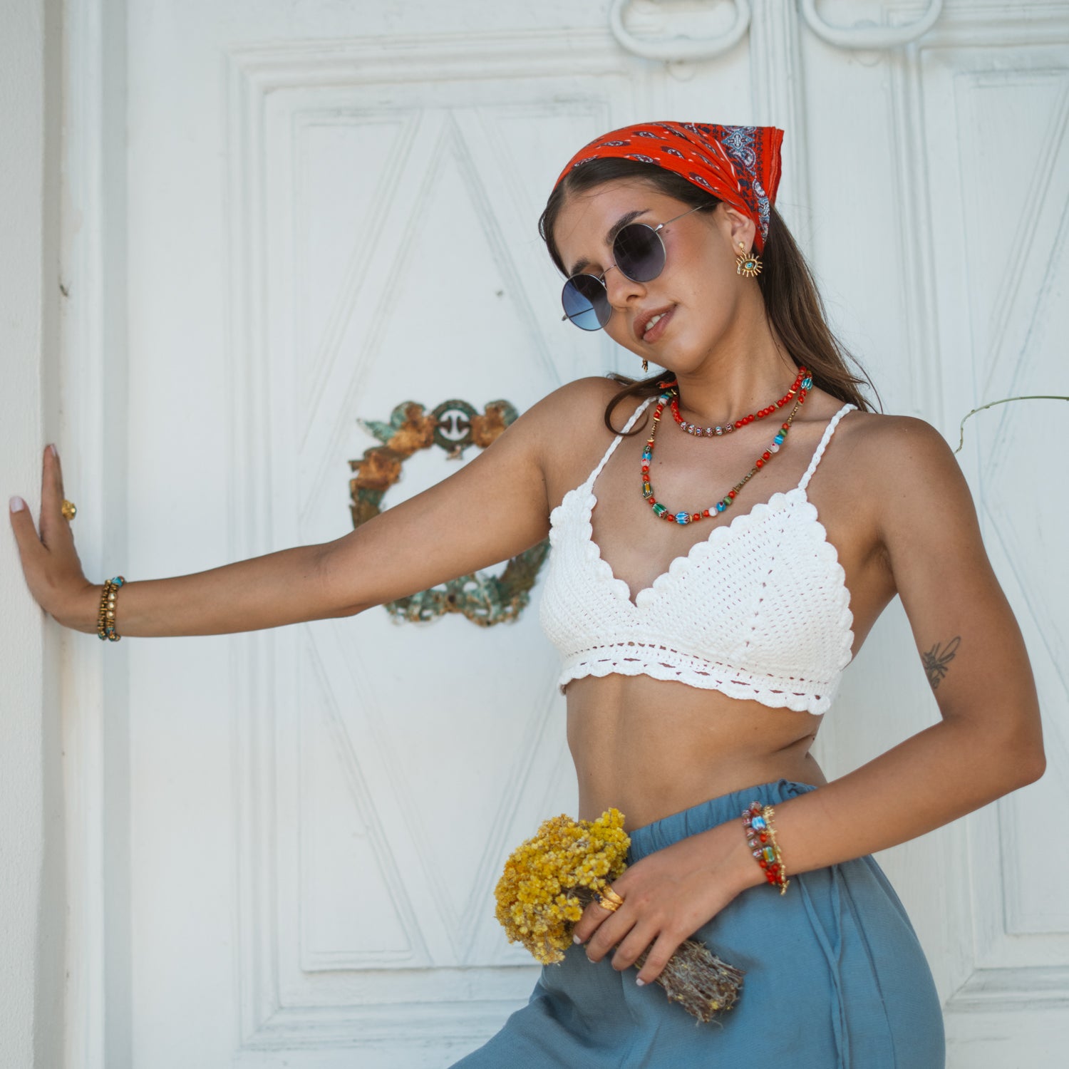 Model leaning against a white door wearing the colorful boho Carnelian Rainbow Energy Beaded Necklace, a white crochet top, and blue linen pants