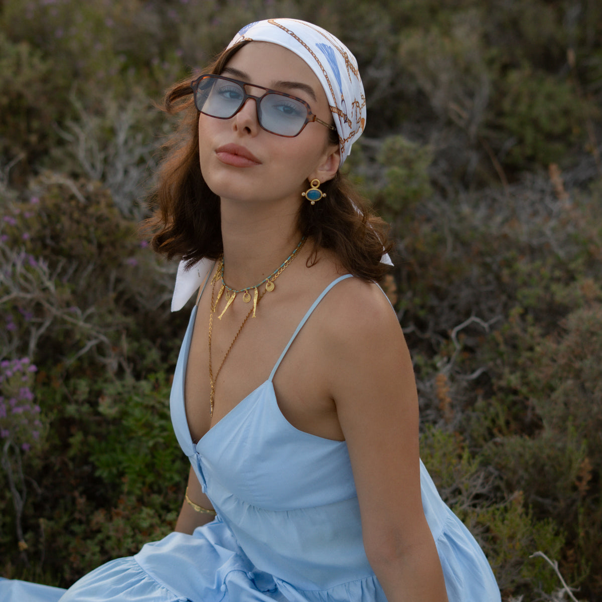Model in a pale blue dress and headscarf outdoors, the blue cat's eye studs catching the light at her ear