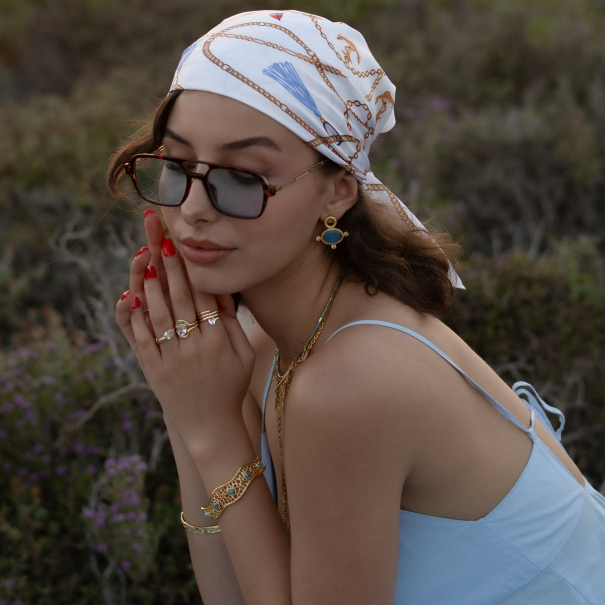Woman resting her chin on her hand, wearing the Cleopatra blue cat's eye studs with a printed headscarf and gold rings