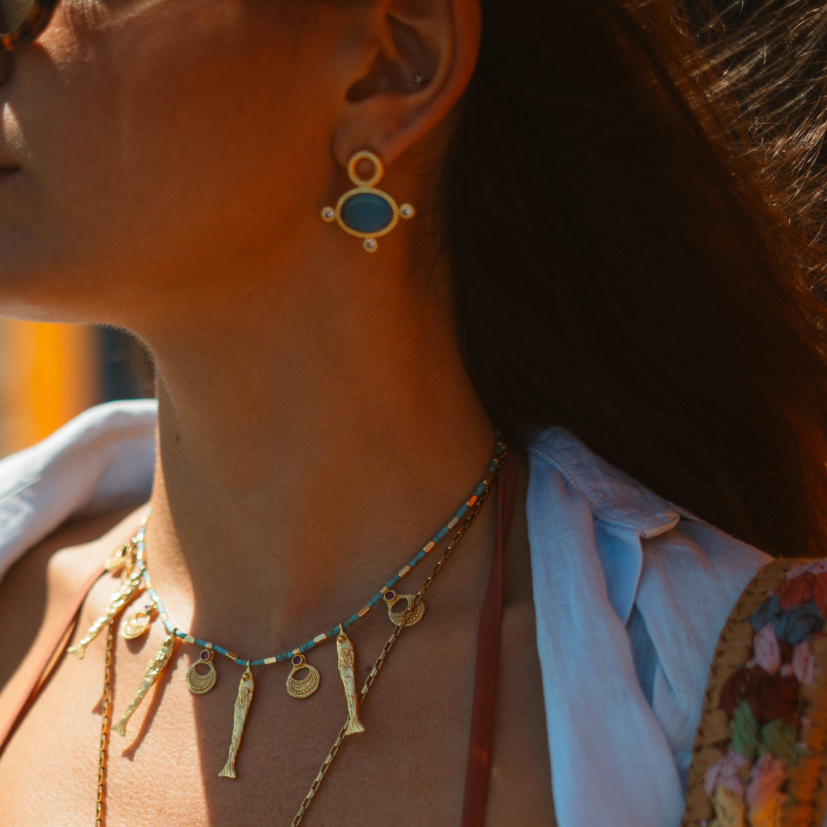 The blue cat's eye gold stud earring worn with a turquoise beaded charm necklace against sunlit skin