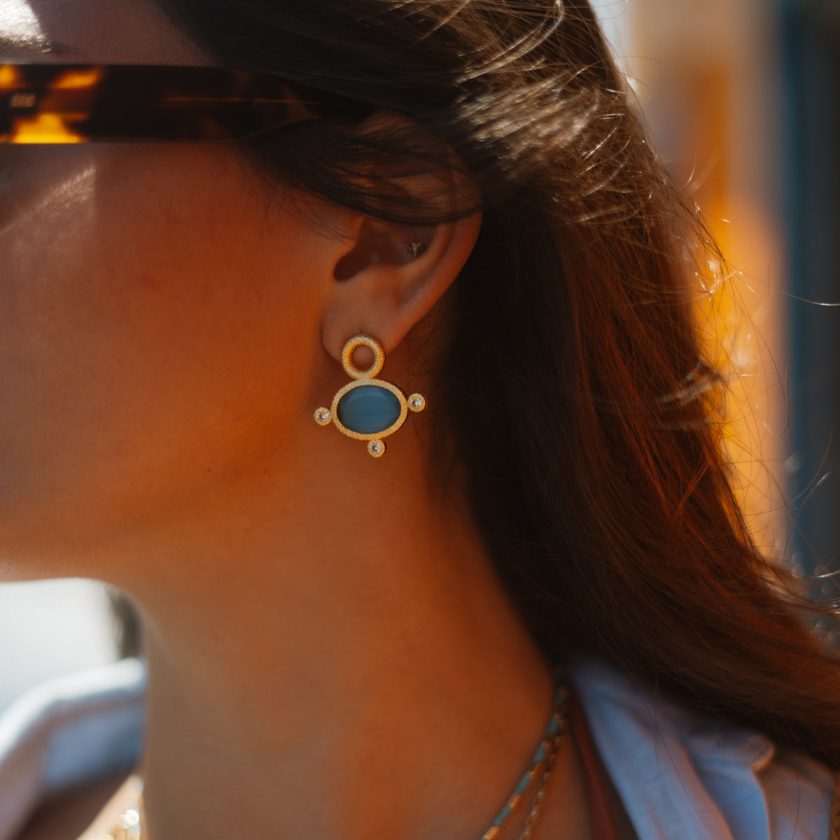 Close up of the blue cat's eye gold stud earring worn on the ear in warm sunlight, hair swept back