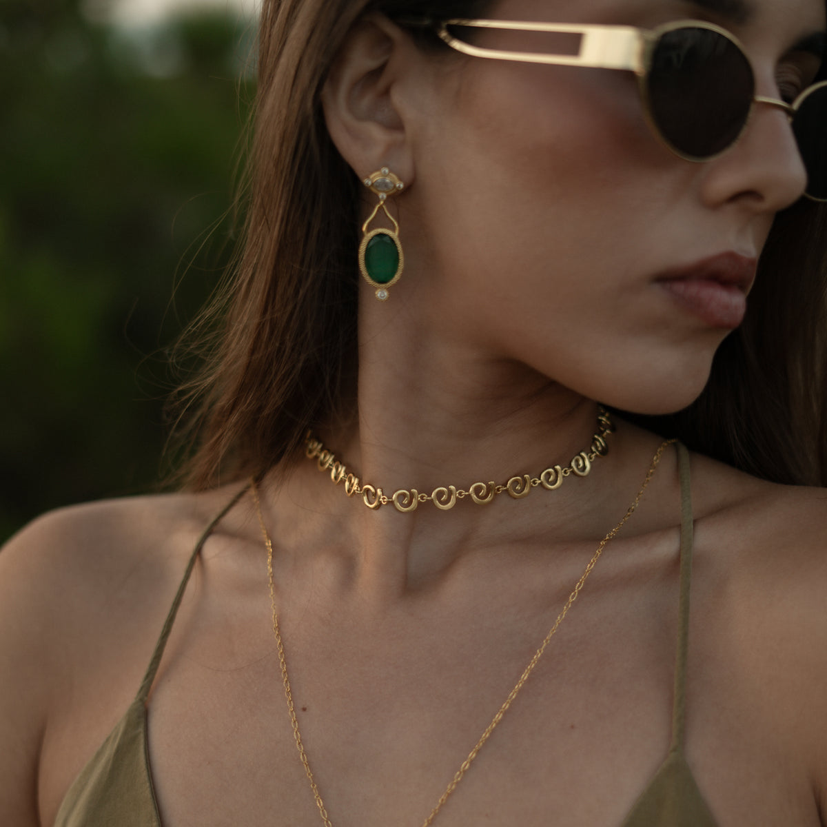 Model in gold sunglasses wearing the Cleopatra Emerald drop earring with a gold spiral link choker