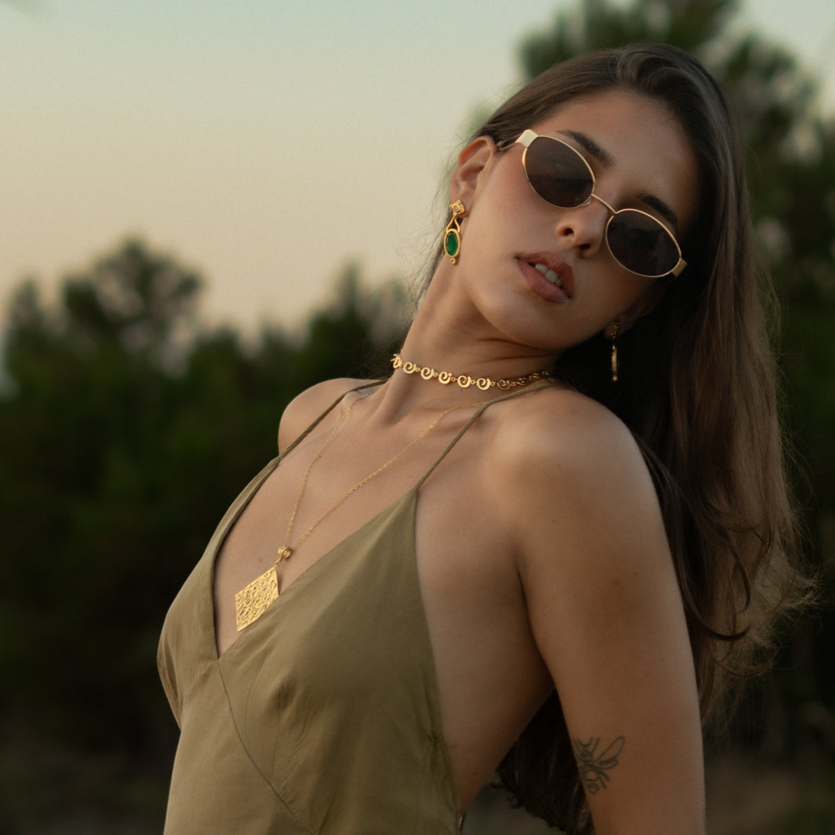 Model in an olive slip dress and sunglasses at dusk, wearing the Cleopatra Emerald cat eye gold drop earrings with a gold choker