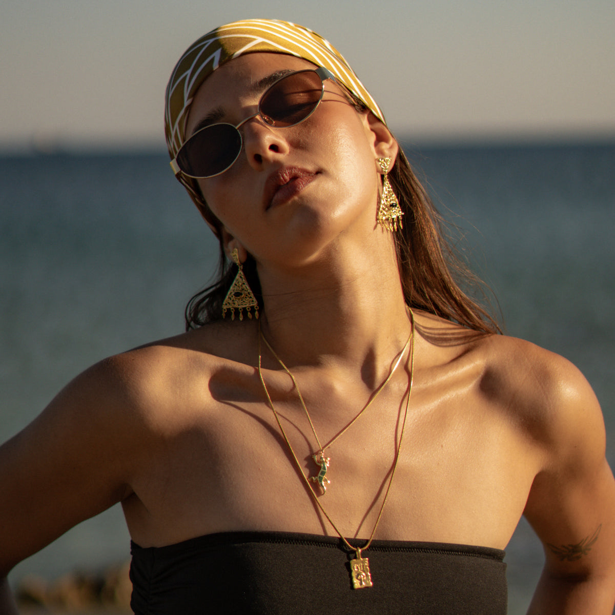 Woman in a silk headscarf and sunglasses by the sea wearing the Cleopatra filigree chandelier earrings with layered gold pendants