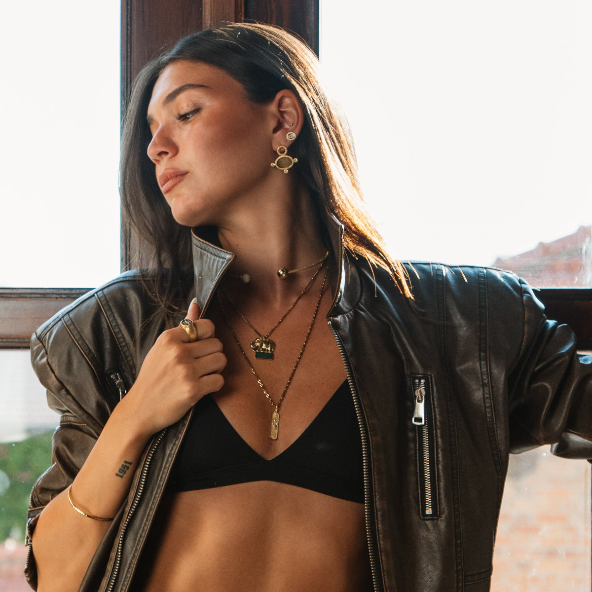 Model holding the collar of a leather jacket, wearing the Cleopatra Golden Citrine cat eye earring and a gold elephant pendant
