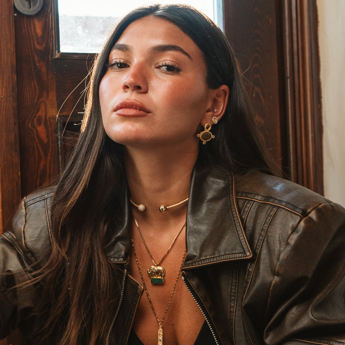 Model in a worn brown leather jacket by a wooden door, wearing the Cleopatra Golden Citrine cat eye earring and layered gold necklaces