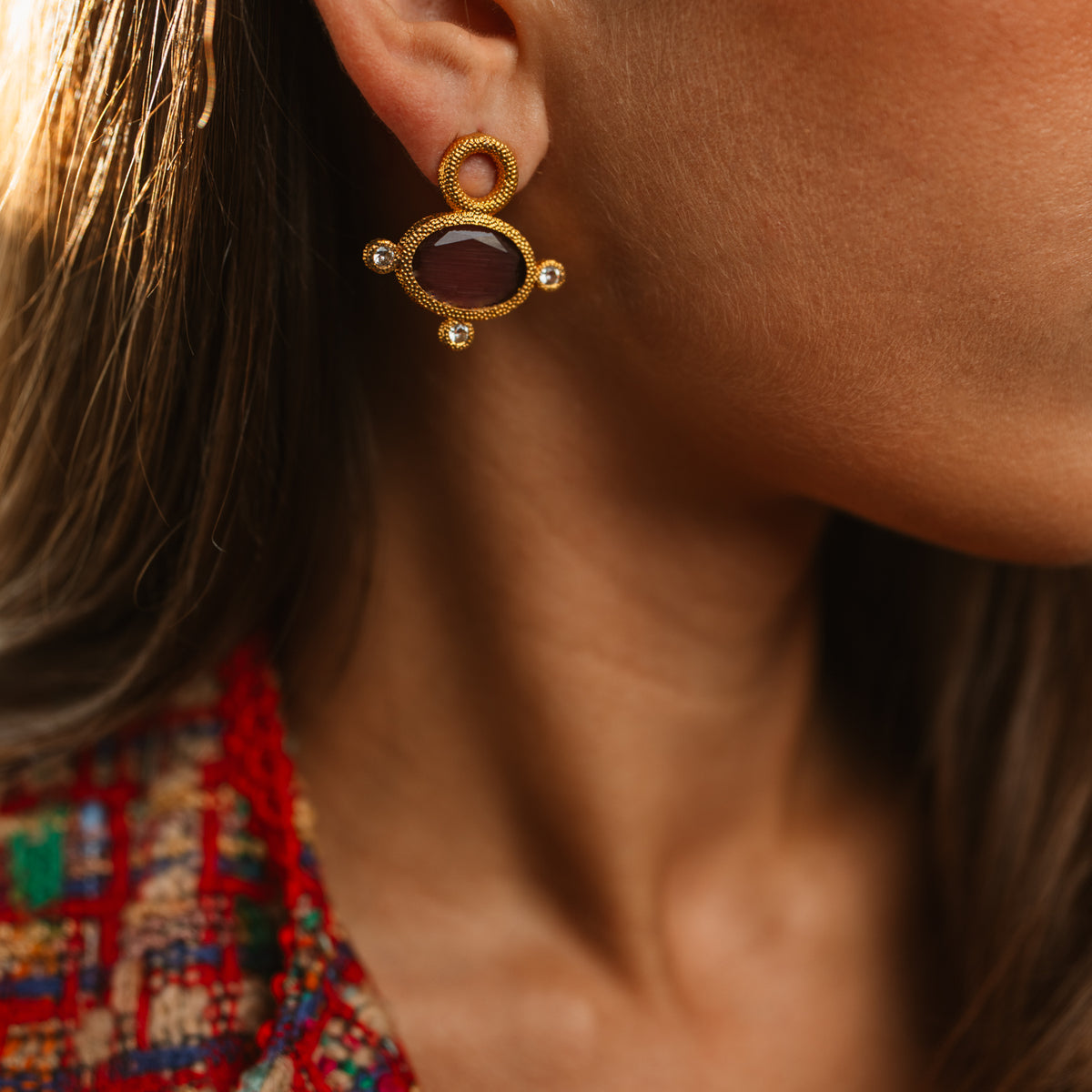 Close view of the Cleopatra Mocha earring on the ear in warm light, the cat eye stone reading deep plum brown above a red tweed collar