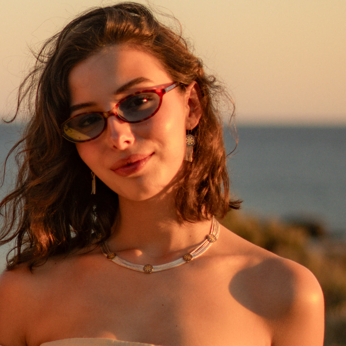 Model by the sea in sunglasses wearing the Cleopatra Mystic two tone necklace and matching tassel earrings