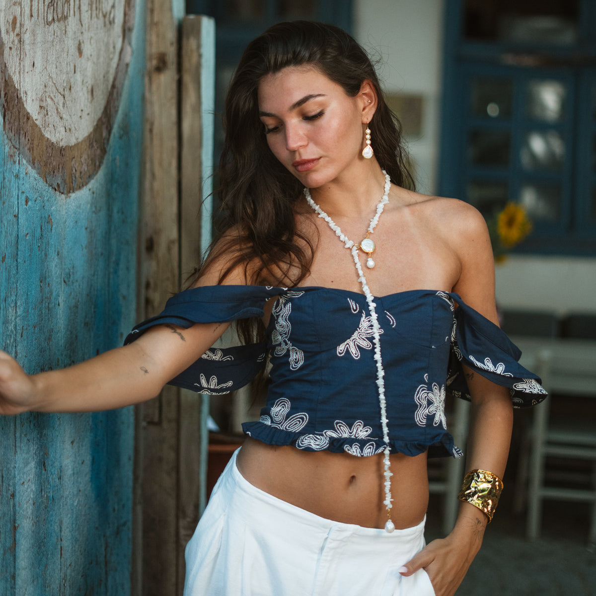 Model leaning on a weathered blue door wearing the Cleopatra pearl drop earrings, a white beaded lariat and a gold cuff
