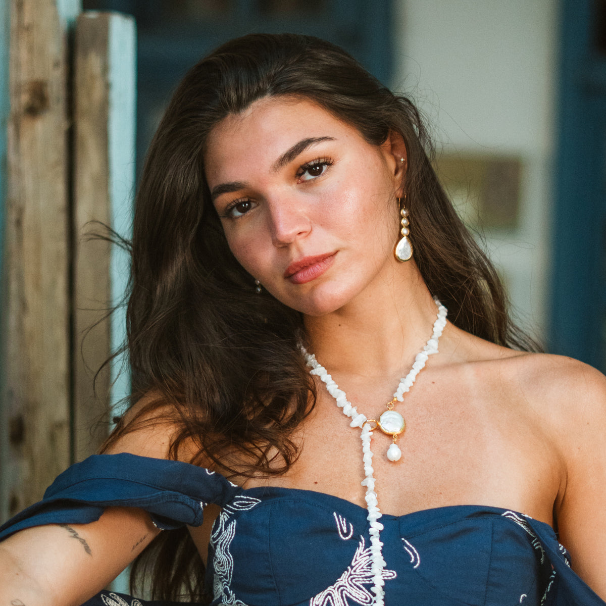 Portrait of a model with long dark hair wearing the pearl cascade gold drop earring and a white beaded necklace with a pearl pendant