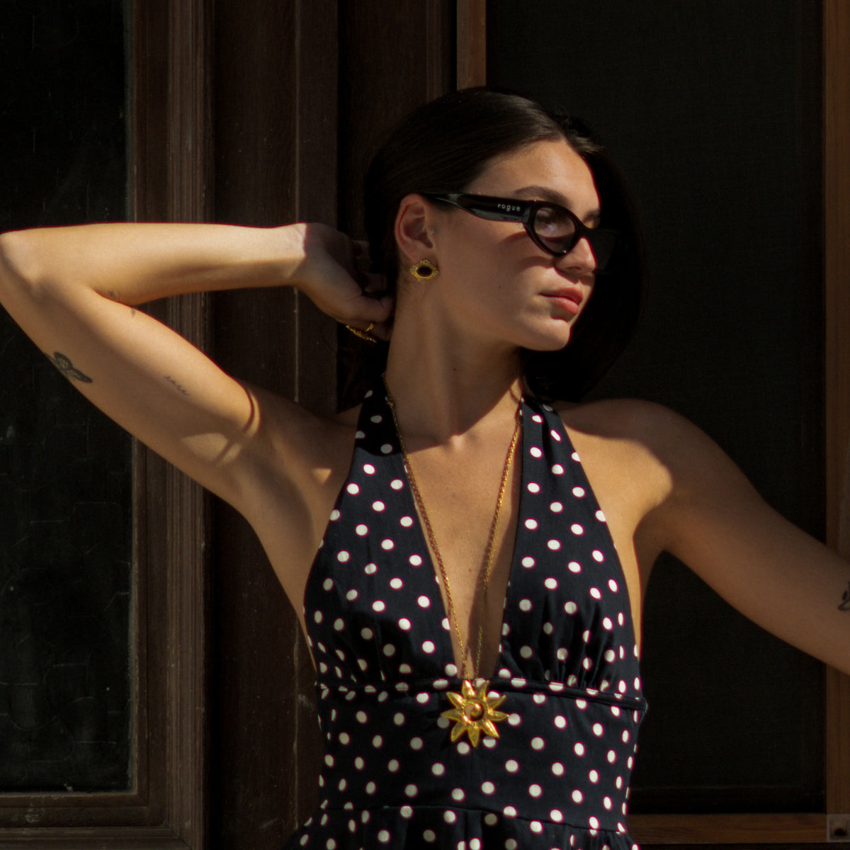 Model in a black polka dot dress wearing the black cat's eye gold studs with a gold sunburst pendant necklace