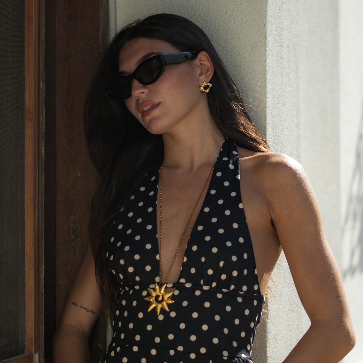 Model leaning against a sunlit wall wearing black cat's eye gold stud earrings and a gold sunburst necklace