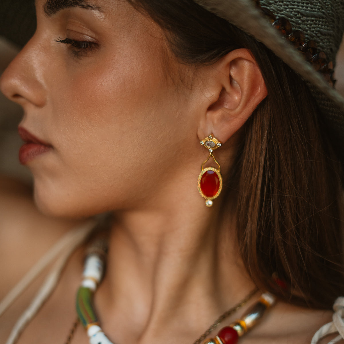 Profile of a model in a straw hat wearing the Cleopatra Sunset drop earring, the orange cat eye turning red in shade