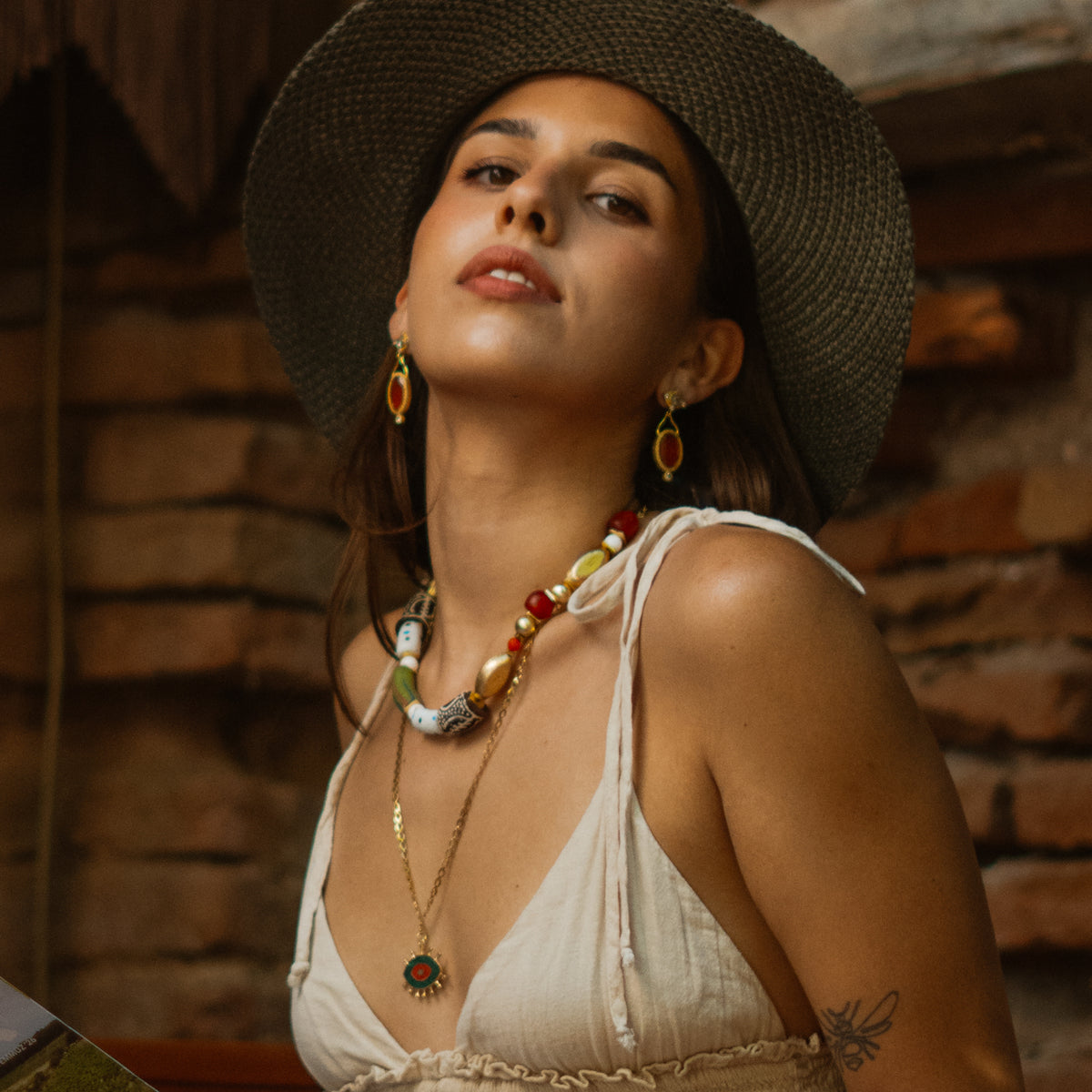 Model in a wide straw hat and beaded necklace wearing the Cleopatra Sunset cat eye gold drop earrings