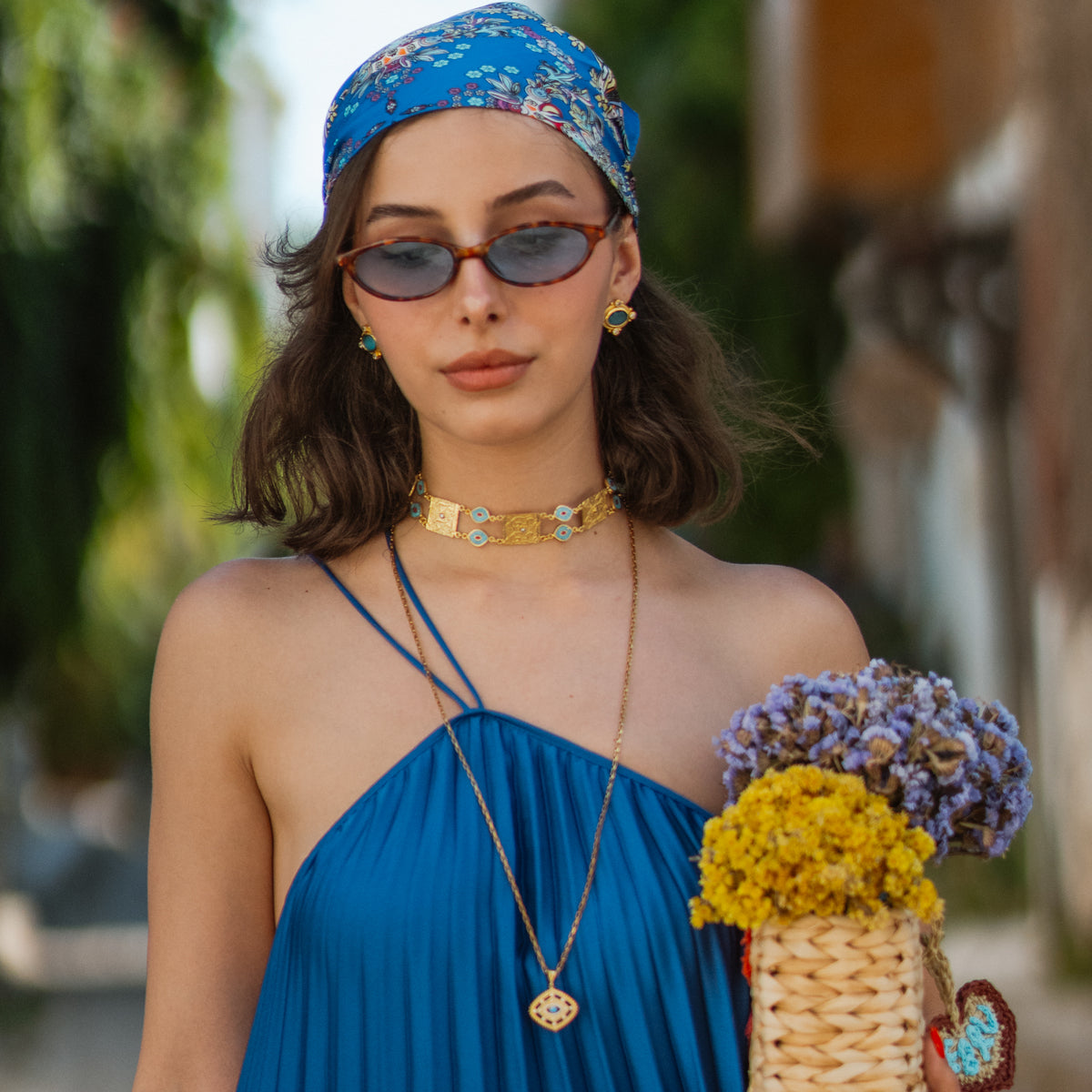 Model in a pleated blue halter dress holding dried flowers, wearing the Cleopatra Turquoise stud earrings and a gold choker