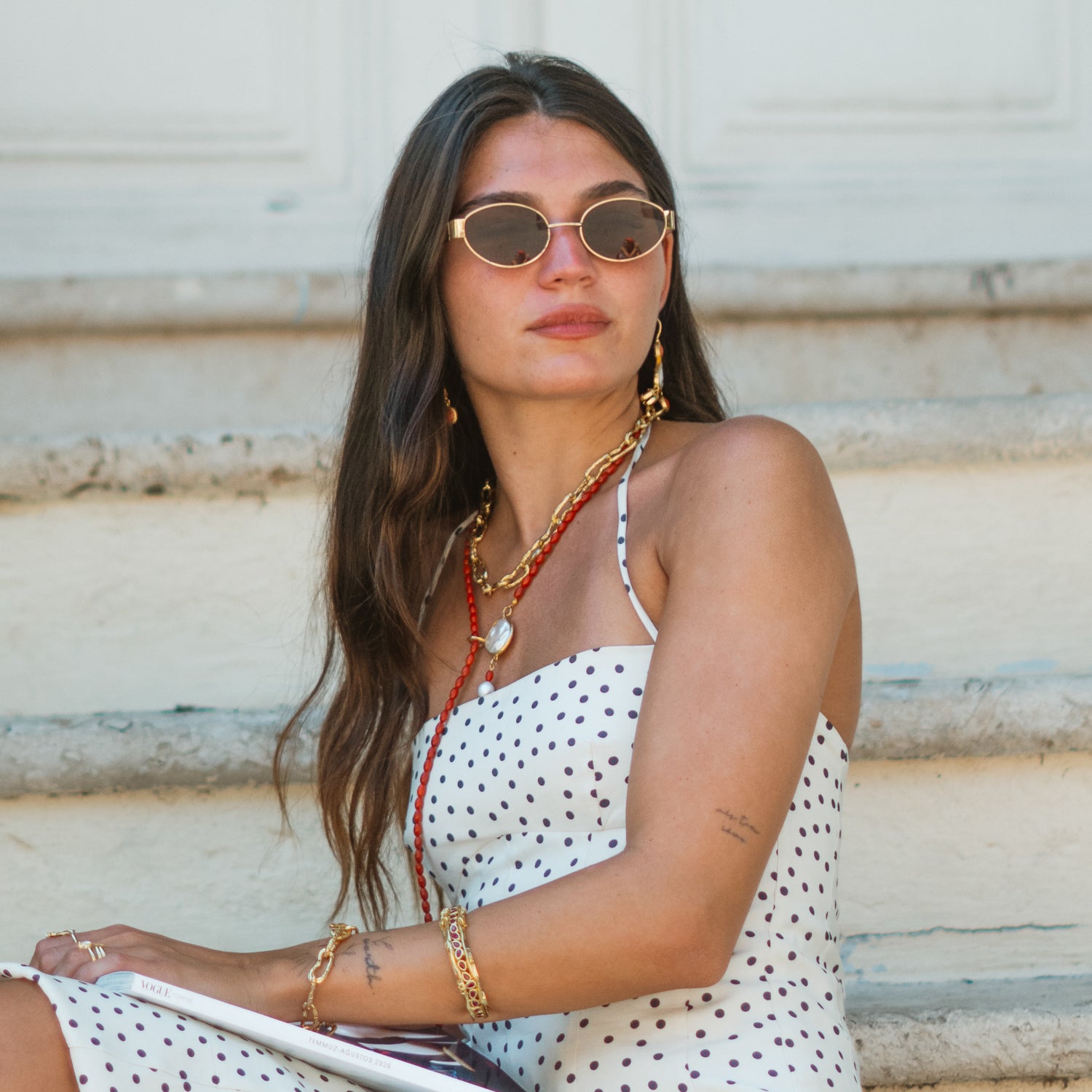 Model wearing the coral pearl lariat necklace layered with a gold chain, seated in a polka dot dress with sunglasses