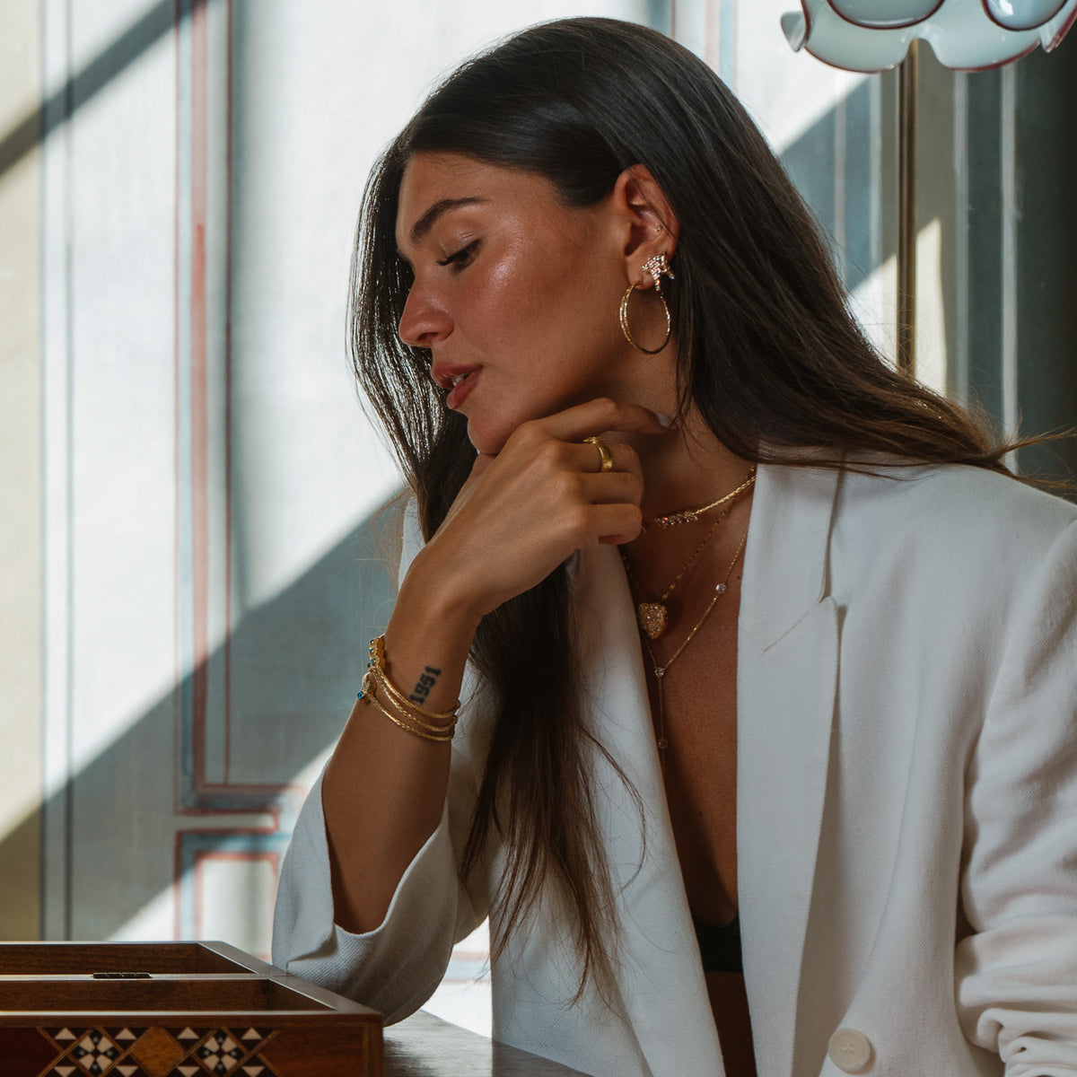Model in a white blazer seated by a window with her hand at her chin, wearing the shooting star stud earrings with layered gold necklaces and bangles