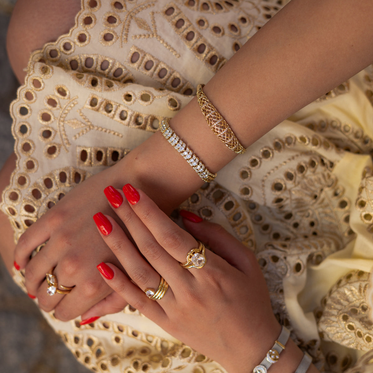 Hands on a cream eyelet dress wearing the gold lattice bangle stacked with a sparkling tennis bracelet and rings