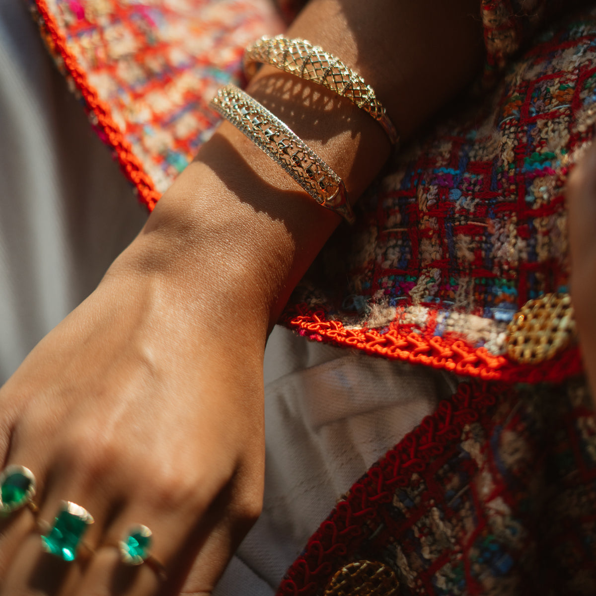 Diamond lattice gold bangle on a sunlit wrist with green stone rings and a tweed jacket