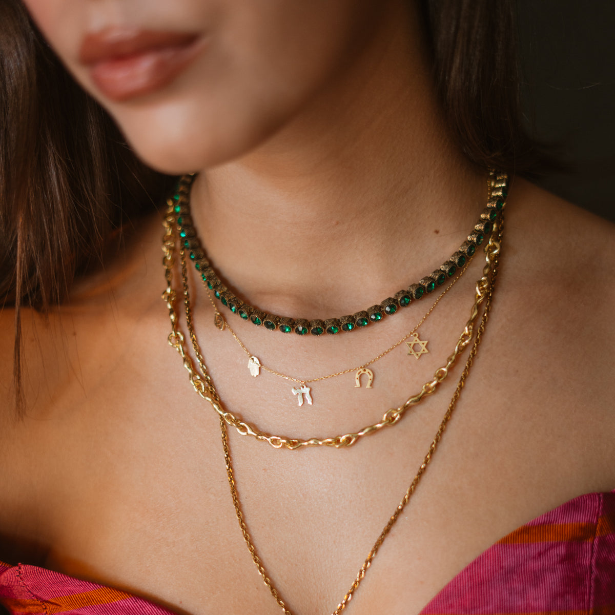 The emerald green tennis necklace worn at the collarbone, layered with a gold charm necklace and a chunky gold chain