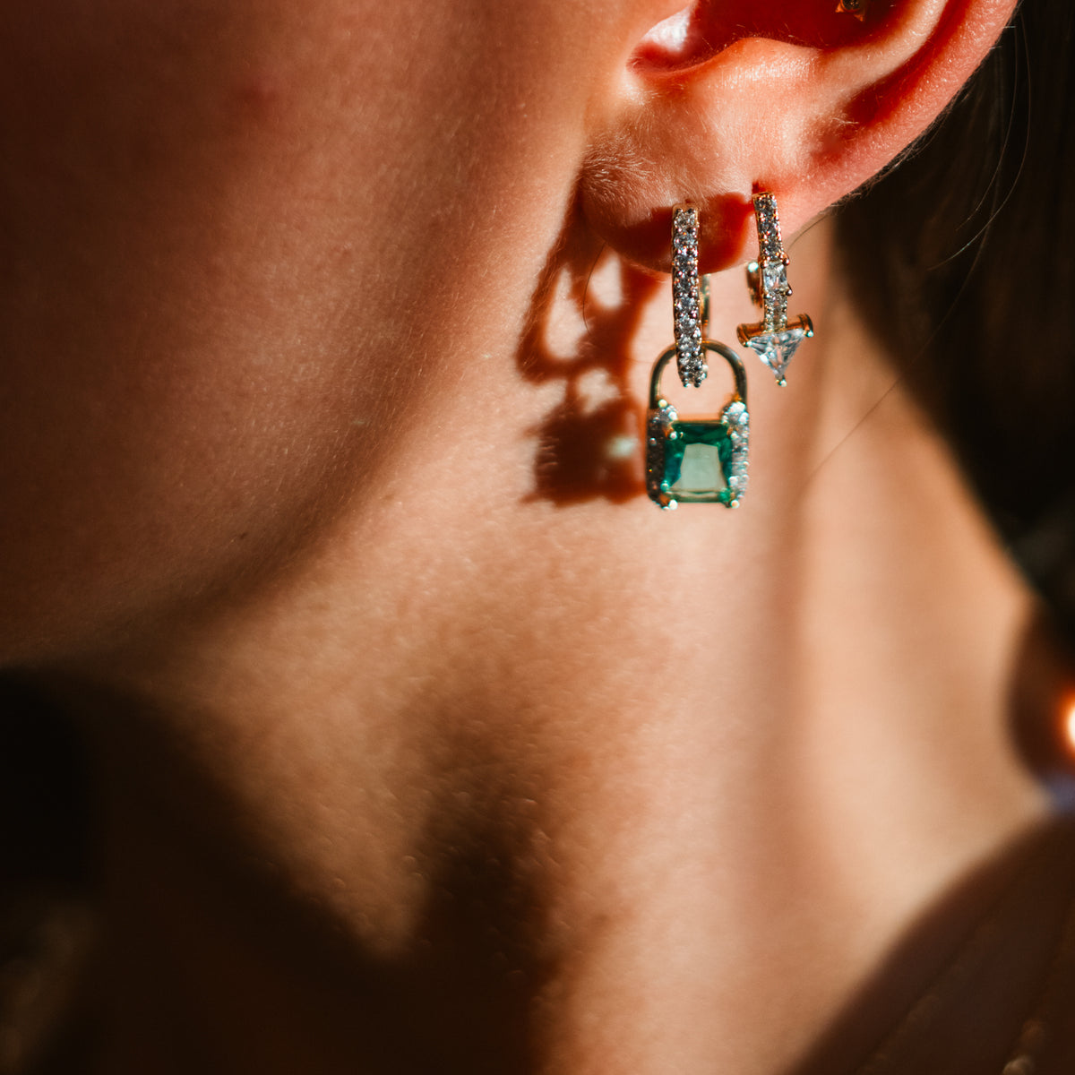 Close up of a mint green zirconia padlock drop huggie earring on the ear, stacked with a second pave huggie in warm light