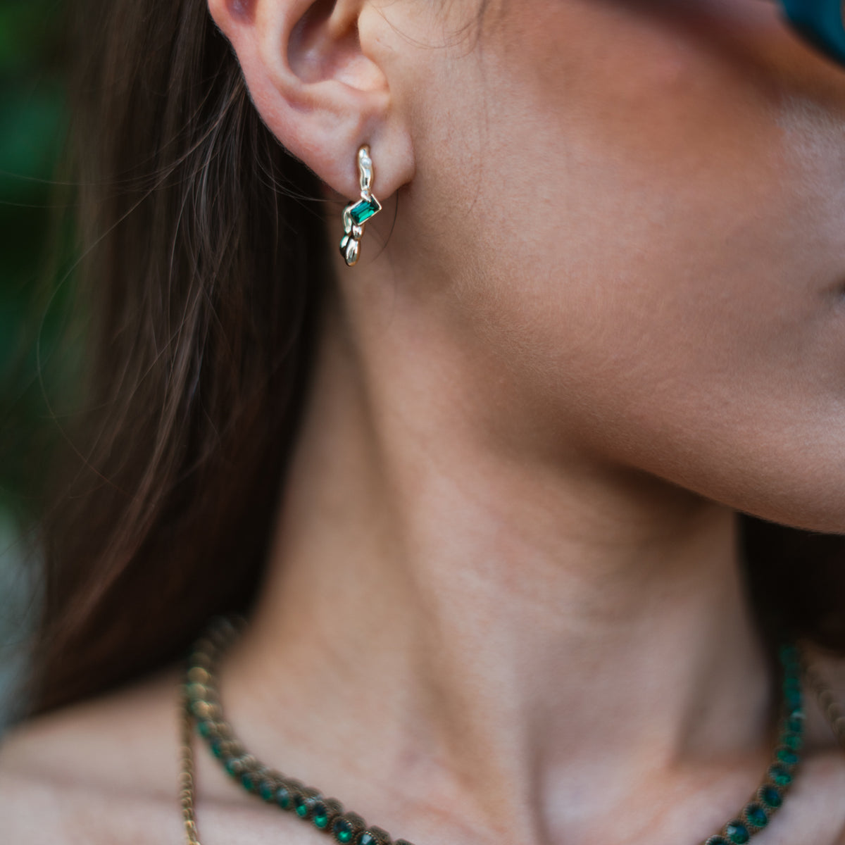 Close view of the Emerald Flow gold earring on a model's ear, worn with a green stone tennis necklace