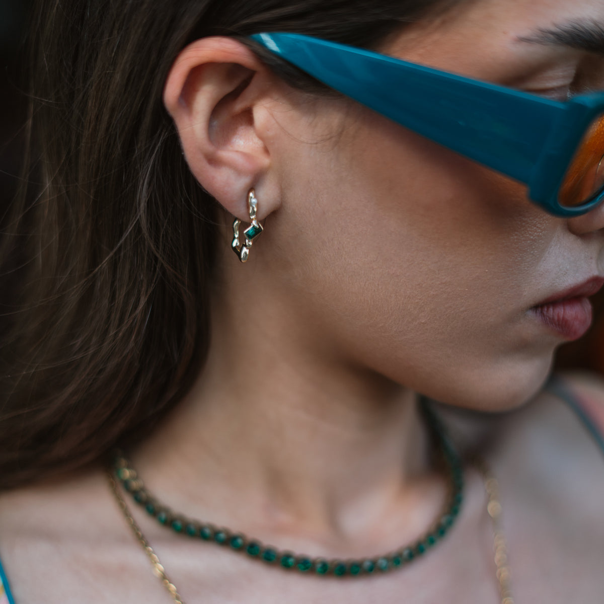 Side profile of a model in teal sunglasses wearing the Emerald Flow gold earring above a green tennis necklace