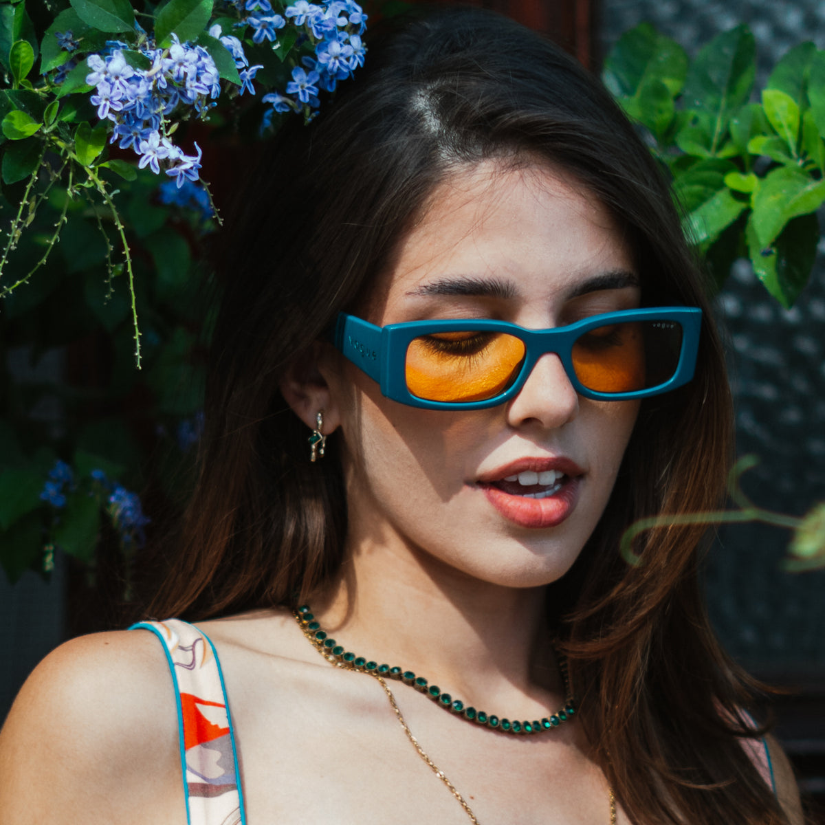 Model in blue sunglasses among blue flowers and green leaves, wearing the Emerald Flow earrings and a green tennis necklace