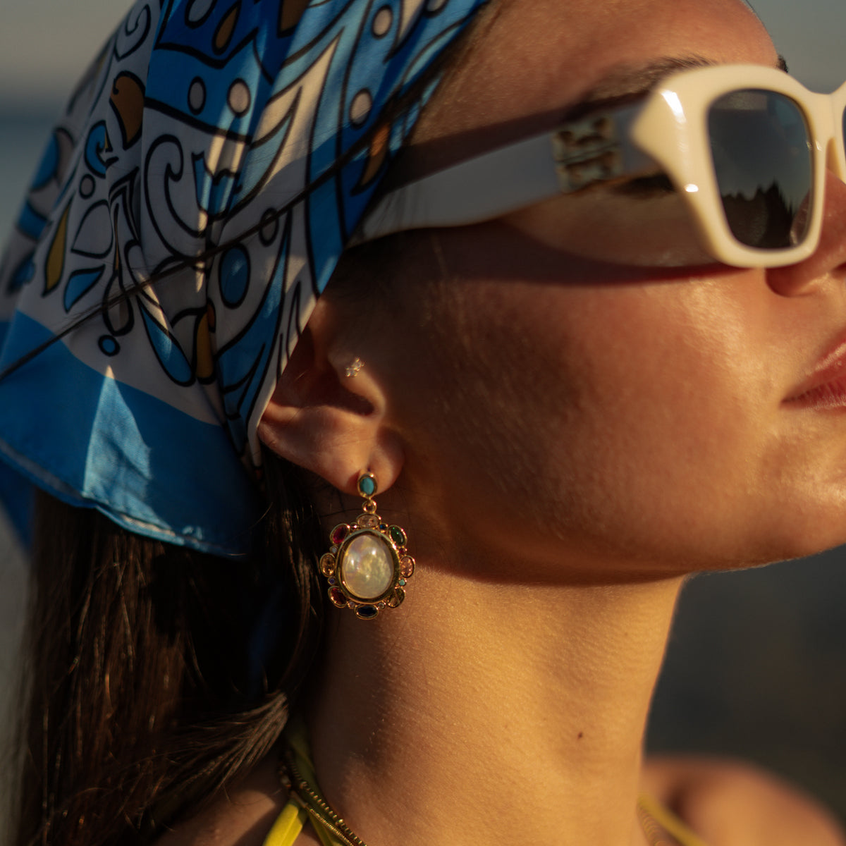 Model looking out to sea wearing baroque pearl multicolor gemstone gold drop earrings and a gold coin pendant necklace