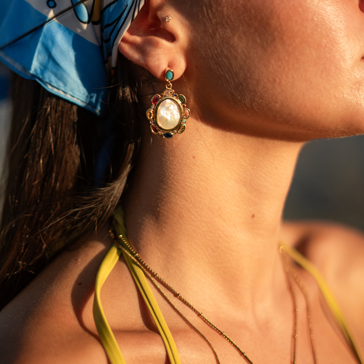 Close view of a baroque pearl and multicolor gemstone gold drop earring worn at the ear in evening light
