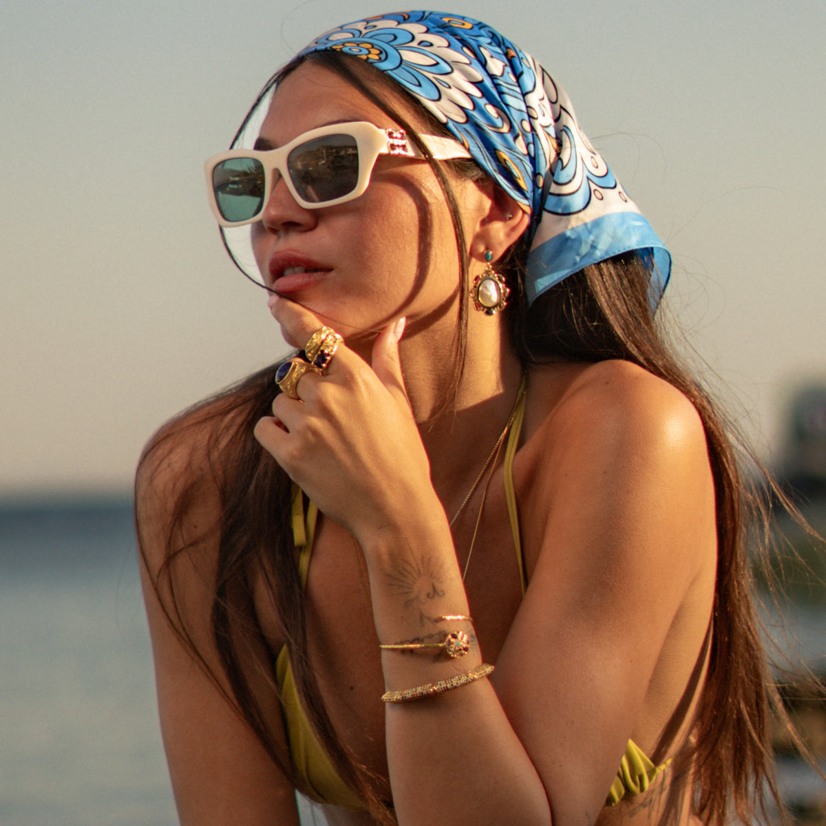 Model by the sea in a blue headscarf wearing baroque pearl multicolor gemstone gold drop earrings with gold rings and bracelets