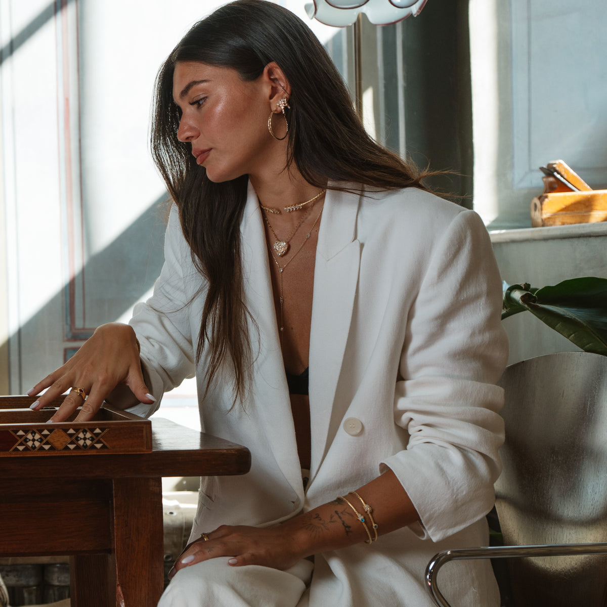 Model in a white suit seated by a sunlit window at an inlaid wooden table, wearing the gold twist hoop earrings with layered necklaces and gold bangles