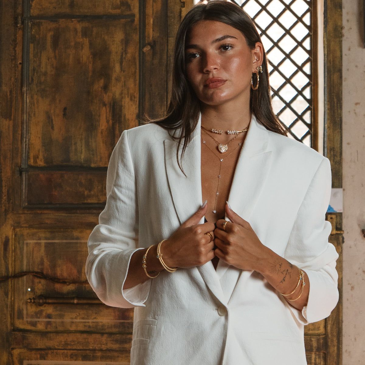 Model in a white blazer wearing the gold twist hoop earrings with layered gold necklaces, bangles and rings