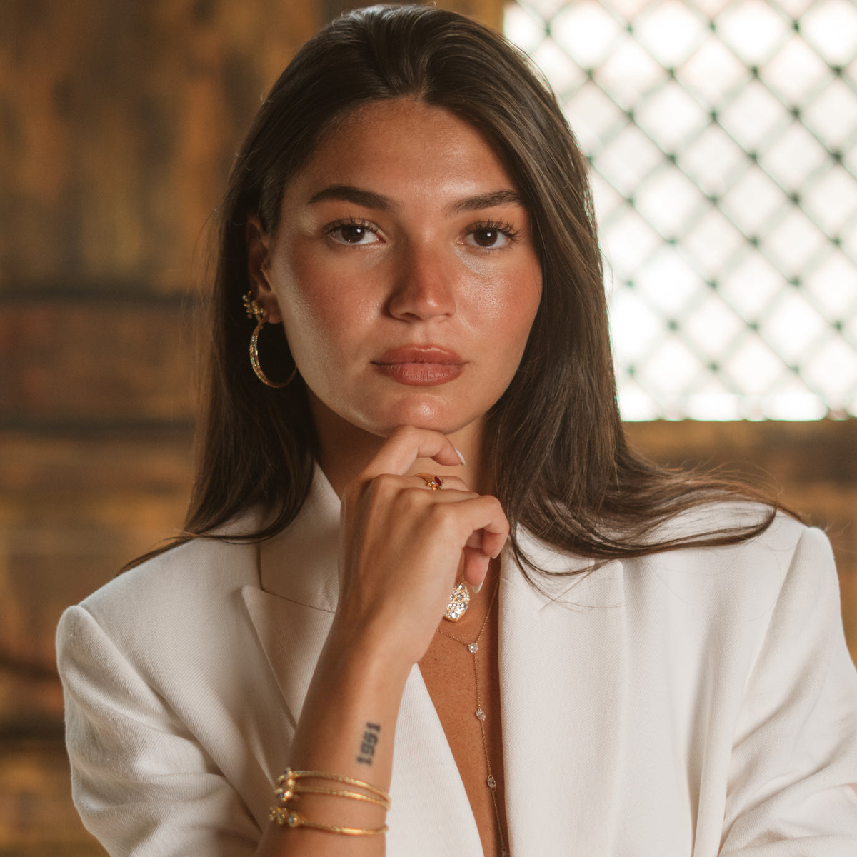 Model resting her hand near her chin, the gold twist hoop earring visible with layered chain necklaces and gold bangles